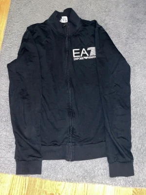 Svart hoodie från Emporio Armani EA7 - Svart hoodie från Emporio Armani EA7 med dragkedja framtill och logga i vitt på bröstet. Klassisk modell med hög krage och långa ärmar. Perfekt för en stilren och sportig look.