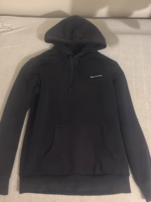 Marinblå hoodie från Peak Performance - Marinblå hoodie från Peak Performance med klassisk huva och dragsko. Stor känguruficka framtill och diskret logga på bröstet. Perfekt för en avslappnad och stilren look. Tillverkad i mjukt material för extra komfort.
