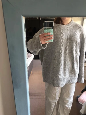 Grå kabelstickad oversized tröja - Mysig grå stickad tröja med kabelstickat mönster framtill och ribbade muddar vid ärmslut och nederkant. Tröjan har en avslappnad, oversized passform och rund halsringning. Perfekt för lager-på-lager och chill dagar.
