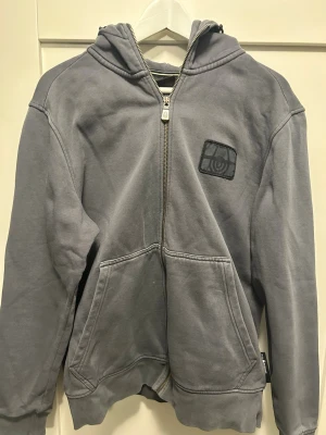 Grå hoodie från Sail Racing - Snygg grå hoodie från Sail Racing med dragkedja framtill, huva och kängurufickor. Diskret logga på bröstet och mjukt material som känns skönt mot huden. Perfekt för en avslappnad och sportig stil.