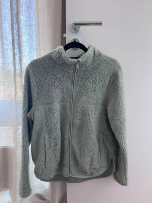 Ljusgrön teddyjacka med dragkedja - Mysig ljusgrön teddyjacka med hel dragkedja framtill och två fickor. Jackan har hög krage och är tillverkad i fluffigt teddy-material som ger en avslappnad och trendig look. Perfekt för lager-på-lager och chill dagar.