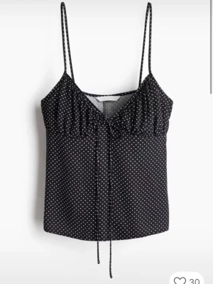 Svart prickig linnetopp med knyt - Svart linnetopp med vita prickar och smala axelband. Toppen har v-ringning och en söt knyt-detalj framtill. Perfekt för varma dagar och enkel att styla med jeans eller kjol.