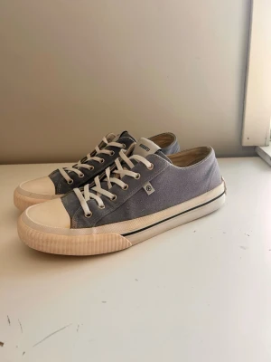 Blåa canvas sneakers från Axel Arigato - Snygga blåa låga sneakers från Axel Arigato i canvas med vita breda skosnören och vit gummisula. Klassisk design med diskret logga på plösen och sidan. Perfekta för en clean och avslappnad stil.