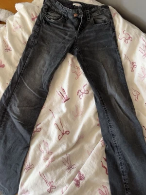 Svarta jeans med rosett-detaljer - Snygga svarta jeans med raka ben och coola rosettbroderier på bakfickorna. Jeansen har klassisk femficksdesign och kopparfärgade nitar. Perfekta för dig som vill ha en unik touch på din outfit.
