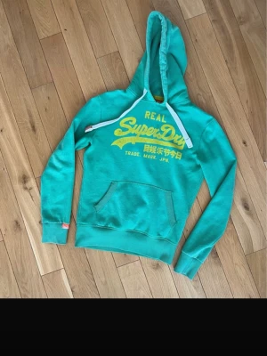 Turkos hoodie från Superdry - Turkos hoodie från Superdry med gul logga och text på bröstet. Klassisk känguruficka framtill, vita dragsnören i huvan och ribbade muddar vid ärmslut och nederkant. Perfekt för en avslappnad streetstyle-look.
