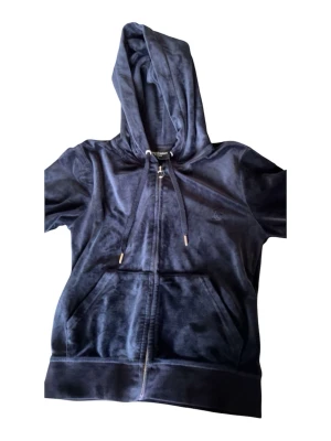 Mörkblå velour hoodie Juicy Couture - Mörkblå hoodie i mjuk velour från Juicy Couture. Dragkedja framtill, huva med dragsko och klassiska kängurufickor. Perfekt för en chill och trendig look. Liten broderad logga på bröstet och ribbade muddar vid ärmslut och nederkant.