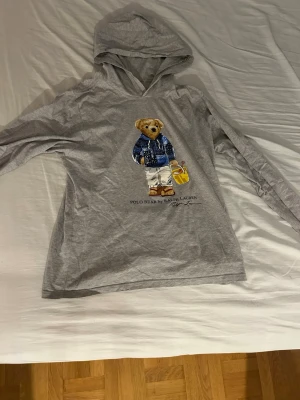 Grå hoodie Polo Bear Ralph Lauren - Grå hoodie från Ralph Lauren med Polo Bear-tryck framtill. Hoodien har långa ärmar och en klassisk huva. Materialet är mjukt och skönt, perfekt för en avslappnad stil. Trycket visar en nallebjörn i blå stickad tröja och vita byxor.