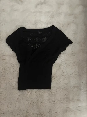 Svart spetsblus med v-ringning - Snygg svart blus med v-ringning och spetsdetaljer över axlar och bröst. Blusen har korta ärmar och är figurnära i midjan. Perfekt för dig som gillar en feminin och stilren look.