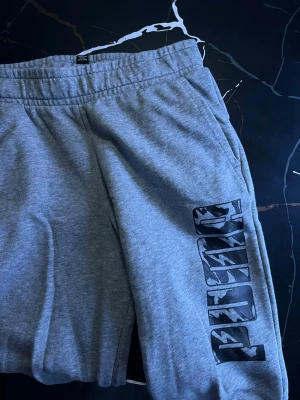Gråa mjukisbyxor från Puma - Snygga grå mjukisbyxor från Puma med svart logga längs benet. Byxorna har resår i midjan och sidofickor. Tillverkade i mjukt och bekvämt material, perfekta för en avslappnad stil. Pris kan sänkas!!