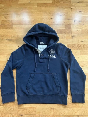  vintage hoodie Abercrombie & Fitch med knappar - Mörkblå hoodie från Abercrombie & Fitch med broderad logga och siffran 1892 på bröstet. Klassisk känguruficka framtill, huva med dragsko och ribbade muddar. Står i storlek L men sitter som M. Skriv om ni har några mer funderingar:)