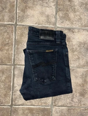 Svarta nudie jeans - Säljer ett par svarta jeans från Nudie  med klassisk femficksdesign och diskret broderad bakficka. Jeansen är i modellen Skinny Lin och har passformen skinny/slim. Storleken är W30L32. Priset är inte hugget i sten, så tveka inte med att skicka prisförslag!