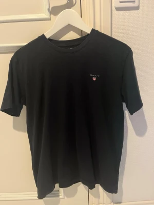 Svart t-shirt från GANT - Klassisk svart t-shirt från GANT med liten logga och sköld på bröstet. T-shirten har rund halsringning och korta ärmar. Perfekt basplagg i mjuk bomull som funkar till allt.