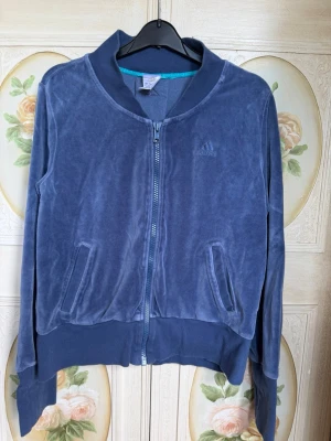 Blå velour fleece från Adidas - Mysig blå fleece i velour från Adidas med dragkedja framtill, ribbade muddar och två fickor. Diskret Adidas-logga på bröstet och snygg detalj med slits bak. 