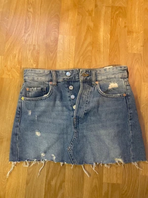Blå jeanskjol med rå kant från H&M - Snygg blå jeanskjol från HM med slitna detaljer, rå kant nertill och klassiska fem fickor. Kjolen har knappar framtill och en cool, avslappnad vibe. Perfekt för dig som gillar en trendig och avslappnad stil.