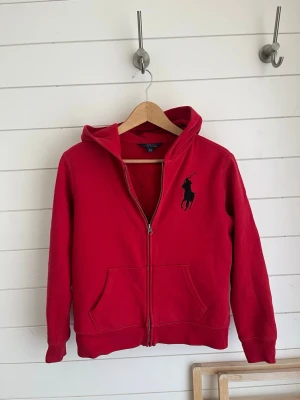 Ralph Lauren Hoodie - Jag säljer en röd Polo Ralph Lauren Hoodie med Dragkedja, tröjan har det klassiska hästemblemet på bröstet men även skriften ”Polo Ralph Lauren” på höger arm. Plagget är gjort i bomull. Tröjan är sparsamt använd och har visuella tecken på användning, I kombination med att nypris ligger på runt 800kr tycker jag att prissättningen är rimlig.