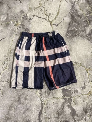 Randiga shorts med marinblå och vita rutor - Snygga shorts med breda marinblå och vita ränder samt en rosa rand framtill. De har elastisk midja med snörning och två sidofickor. Perfekta för sommaren och har en sportig vibe. Materialet känns lätt och syntetiskt.