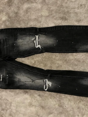 Svarta slitna jeans med hål - Säljer ett par svarta jeans med slitningar och hål på knäna. Jeansen har skinny fit och coola detaljer som blekta partier och små färgstänk. Perfekta för en streetwear-look och passar dig som gillar en edgy stil.