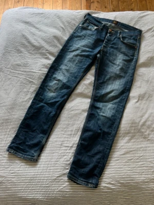 Mörkblå raka jeans från Lee - Snygga mörkblå jeans från Lee med raka ben och klassisk femficksdesign. W31 L32. Jeansen har slitningar och en cool tvätt som ger en avslappnad look. Tillverkade i denim med Lee-logga på bakfickan. Perfekta för en casual stil.
