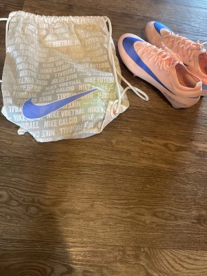 Nike Mercurial rosa/blå fotbollsskor - Säljer ett par Nike Mercurial fotbollsskor i en snygg ljusrosa färg med blå detaljer och Nike-logga. Skorna har låg profil, snörning och är tillverkade i syntetmaterial för bästa grepp och känsla på planen. Medföljer en vit Nike-påse med tryck. Jag köpte dem på unisport.store dem kostade 3300 eller nåt sånt och dem är elite å äkta. Jag har använt dem en gång