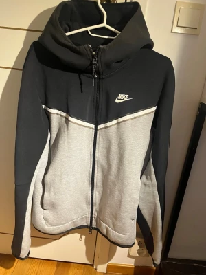 Nike hoodie med dragkedja och huva - Snygg hoodie från Nike i svart och ljusgrått med vit logga på bröstet. Modellen har dragkedja framtill, huva med snören och en cool detalj med ficka på ärmen. Materialet är mjukt och bekvämt, perfekt för chill eller träning.