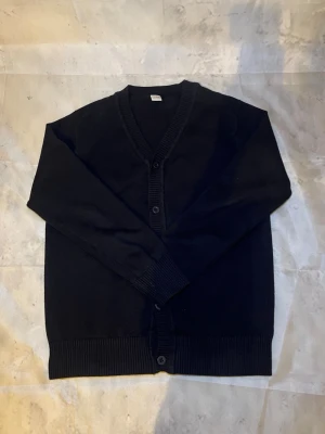 Svart basic kofta med knappar - En enkel svart kofta med v-ringning och tre knappar framtill. Koftan har ribbade muddar vid ärmslut och nederkant, vilket ger en clean och stilren look. Perfekt att slänga över en t-shirt eller skjorta för en avslappnad vibe.