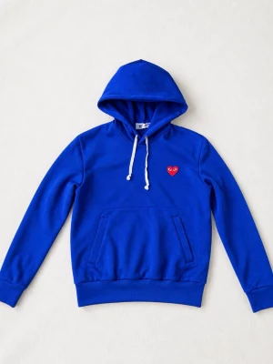 Blå hoodie från Comme des Garçons - Snygg blå hoodie från Comme des Garçons med klassisk huva och vita dragsnören. På bröstet sitter det ikoniska röda hjärtat med ögon. Hoodien har en stor magficka och ribbade muddar vid ärmar och nederkant. Perfekt för en chill och trendig look.