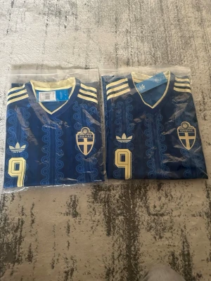 Blå Sverige retro fotbollströja Adidas - Retroinspirerad blå fotbollströja från Adidas med Sveriges landslagsemblem och nummer 9 på bröstet. Tröjan har gula detaljer, klassiska Adidas-ränder på axlarna och ett mönster i tyget. Perfekt för dig som vill sticka ut på match eller träning.