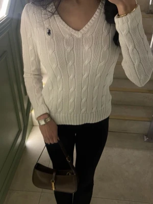 Vit kabelstickad tröja från Ralph Lauren - Vit kabelstickad tröja från Ralph Lauren med V-ringning och klassisk marinblå logga broderad på bröstet. Tröjan har långa ärmar och ribbade muddar vid ärmslut och nederkant. Perfekt för en stilren och tidlös look.