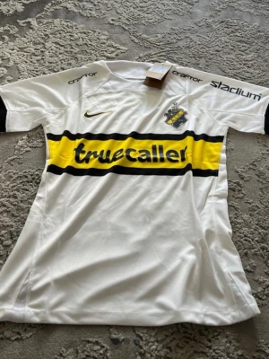 Vit AIK fotbollströja Nike - AIK:s officiella vita fotbollströja från Nike med klubbmärke på bröstet och sponsorlogga 'truecaller' i gult och svart över bröstet. Tröjan har svarta detaljer på ärmarna och tryck på axlarna. Perfekt för match eller träning.