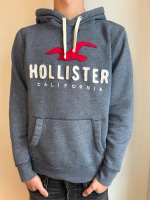 Blå Hollister hoodie med röd logga - Mysig blå hoodie från Hollister med stor vit broderad logga och röd fågel på bröstet. Klassisk känguruficka framtill, vita dragsnören i huvan och mjukt material. Perfekt för en chill och avslappnad stil. Strl: M passar S✅ Pris kan diskuteras!💸 Hör av dig om du har funderingar!👍