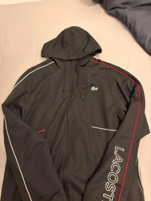 Svart hoodie från Lacoste med tryck - Snygg svart hoodie från Lacoste med dragkedja, huva och dragsko. Framsidan har vita och röda detaljer samt Lacoste-logga på bröstet och stort tryck med texten 'LACOSTE' på ärmen. Baksidan har en stor krokodil i vitt och rött. Perfekt för en sportig och trendig look.
