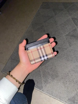 Beige rutigt korthållare Burberry - Snygg korthållare från Burberry i klassiskt beige rutigt mönster med svarta, vita och röda detaljer. Översta kanten är i svart läder vilket ger en lyxig känsla. Perfekt för kort och ID, smidig att ha i fickan.