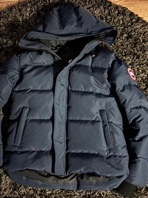 Mörkblå dunjacka från Canada Goose - Snygg mörkblå dunjacka från Canada Goose med huva och klassisk logotyp på ärmen. Jackan har två stora framfickor, dragkedja och vindslå. Perfekt för kalla vinterdagar och riktigt varm tack vare dunfyllningen. M men passar s