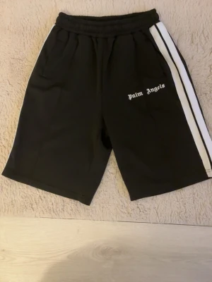 Svarta Palm Angels shorts med vit rand - Svarta shorts från Palm Angels med vit sidrand och logga tryckt på vänster ben. Modellen har elastisk midja och sportig look, perfekta för en avslappnad stil. Storlek m passar L