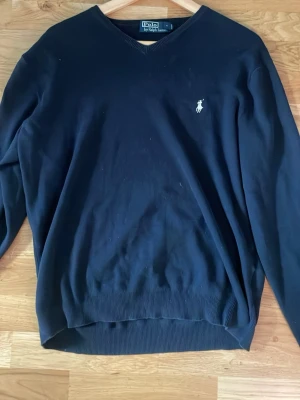 Mörkblå stickad tröja Polo Ralph Lauren - Klassisk mörkblå stickad tröja från Polo Ralph Lauren med V-ringning och den ikoniska vita logon broderad på bröstet. Tröjan har ribbade muddar vid ärmslut och nederkant. Perfekt för en stilren och enkel look.