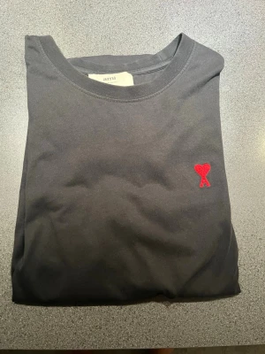 Svart t-shirt från Ami Paris - Svart t-shirt från Ami Paris med klassisk passform och rund hals. På bröstet finns Ami:s ikoniska röda hjärta med bokstaven A broderat. Tillverkad i mjuk bomull som känns skön mot huden. Perfekt för en clean och stilren look. Storlek L men passar M och säljer för endast 350kr. Skriv för fler frågor!
