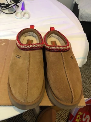 UGG bruna mockatofflor med röd kant - Bruna tofflor från UGG i mjuk mocka med vitt fluffigt foder. Dekorativ röd och vit kant runt öppningen och röd ögla bak. Rejäl sula för extra komfort och värme, perfekta för att chilla hemma.