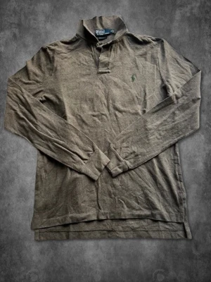 Brun långärmad piké från Polo Ralph Lauren Stl M - Klassisk brun långärmad pikétröja från Polo Ralph Lauren med broderad grön logga på bröstet. Skjortan har krage, knappar vid halsen och är tillverkad i mjuk bomull. Perfekt för en avslappnad men ändå stilren look.