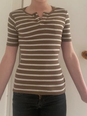 Brun och vit randig ribbad topp - Snygg brun ribbad topp med vita horisontella ränder och korta ärmar. Toppen har en rund halsringning med knappdetalj framtill och sitter tight mot kroppen. Perfekt för dig som gillar en enkel men trendig stil.
