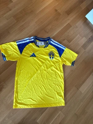 Svensk landslagströja Adidas gul/blå - Säljer en gul svensk landslagströja från Adidas med blå detaljer och vita ränder på axlarna. Tröjan har broderad landslagssköld, Adidas-logga och texten 'Sverige' på ryggen. Tillverkad i lätt och ventilerande material, perfekt för fotboll eller träning.