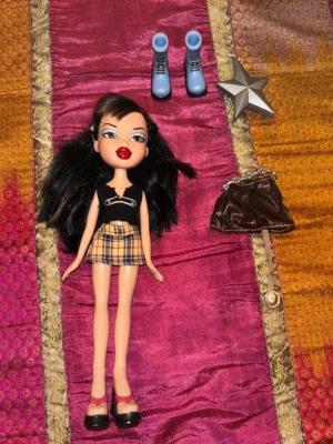Vintage Bratz-docka med plaidkjol – Y2K / tidigt 2000-tal - Original Bratz-docka från tidigt 2000-tal med klassisk Y2K-look och ikonisk Bratz-stil. Dockan har mörkt hår, markerade drag och outfit med rutig kjol, svart topp och skor enligt bilderna.  Extra accessoarer och delar ingår också enligt bilderna, perfekt för samlare eller den som vill komplettera sin Bratz-samling.  Dockan är i mycket bra vintage-skick med välbevarat ansikte, hår och kropp. Kläder och accessoarer är i fint skick och helhetsintrycket är mycket fint.  