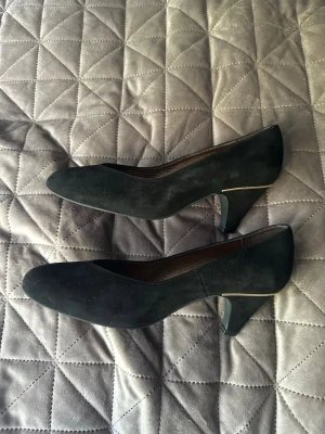 Svarta mocka pumps med låg klack - Stilrena svarta pumps i mocka med låg klack och diskret gulddetalj vid hälen. Klassisk spetsig tå och elegant form som passar till många olika looks. Perfekta för dig som gillar en tidlös och chic stil.