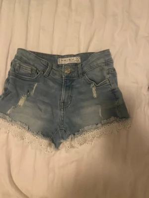 Blå jeansshorts med spetskant - Säljer ett par ljusblå jeansshorts från Amisu med snygga slitningar och vit spetskant längst ner. Shortsen har fem fickor, bälteshällor och knappstängning. Perfekta för sommaren och ger en söt men cool vibe.