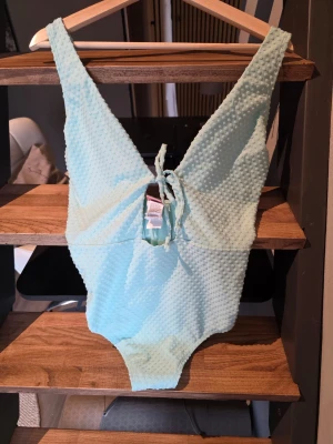 Ljusblå baddräkt med struktur Hunkemöller maui - Ljusblå baddräkt med djup v-ringning och knytning framtill. Materialet har en prickig struktur som ger en cool och unik look. Perfekt för strand eller pool och riktigt snygg att matcha med solglasögon.