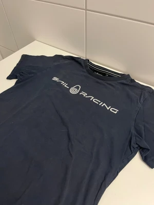 Mörkblå Sail Racing t-shirt - Snygg mörkblå t-shirt från Sail Racing med vit logga tryckt över bröstet. Klassisk rund halsringning och korta ärmar. Perfekt för dig som gillar stilrena och sportiga plagg. Tillverkad i mjuk bomull för skön känsla hela dagen.