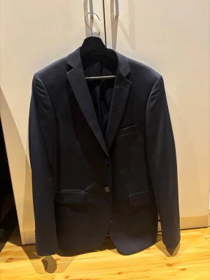 Mörkblå klassisk kavaj - Stilren mörkblå kavaj med två knappar framtill, klassiska slag och tre fickor. Fodrad insida med diskreta sömmar. Perfekt för en dressad look. Materialet ser ut att vara en blandning av polyester och viskos. Storlek 46/M