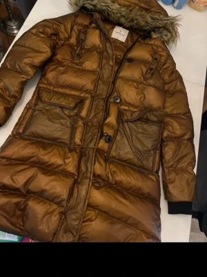 Brun lång dunjacka från Moncler - Snygg brun dunjacka från Moncler med lång passform, stora fickor framtill och huva med fuskpäls. Jackan har dragkedja och knappar, samt ribbade muddar vid ärmsluten. Perfekt för kalla vinterdagar och riktigt varm. Finns en liten reva längst ner därav priset