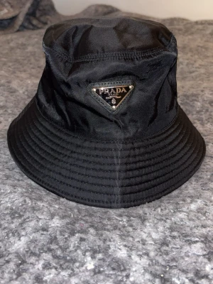 Svart bucket hat från Prada - Säljer en svart bucket hat från Prada med ikonisk metallogo framtill. Hatten är tillverkad i nylon och har en klassisk form med bred, nedåtlutande brätte. Perfekt accessoar för en trendig streetstyle-look.