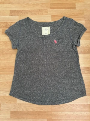 Grå topp från Abercrombie & Fitch - Gråmelerad topp från Abercrombie & Fitch med korta ärmar och rund halsringning. Toppen har knappdetaljer framtill och en liten bröstficka med rosa logga. Mjuk och skön bomullskvalitet, perfekt till jeans eller kjol. Lite nopprig, men syns inte pga färg grå som blandad med lite vit, därav billigt pris på 70kr. Pok.
