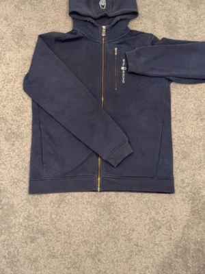 Marinblå hoodie från Sail Racing - Snygg marinblå hoodie från Sail Racing med dragkedja framtill och broderad logga på bröstet. Hoodien har en huva och en praktisk bröstficka med dragkedja. Tillverkad i mjukt bomullsmaterial, perfekt för en avslappnad stil. Färgen på hoodien är en aning blekt, men fortfarande riktig tjusig.